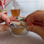 Deux mains ajustent une charlotte alimentaire lavable à feuilles de palmier sur un petit ramequin en verre contenant une préparation jaune, son étiquette de marque et un ruban tricolore étant visibles.
