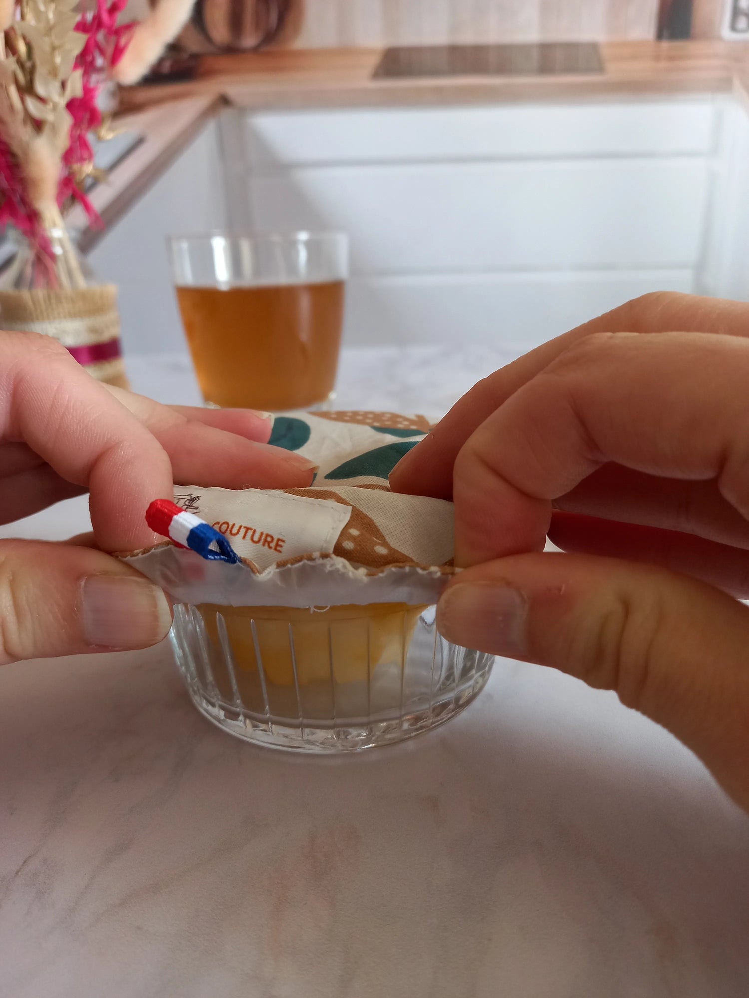 Deux mains ajustent une charlotte alimentaire lavable à feuilles de palmier sur un petit ramequin en verre contenant une préparation jaune, son étiquette de marque et un ruban tricolore étant visibles.