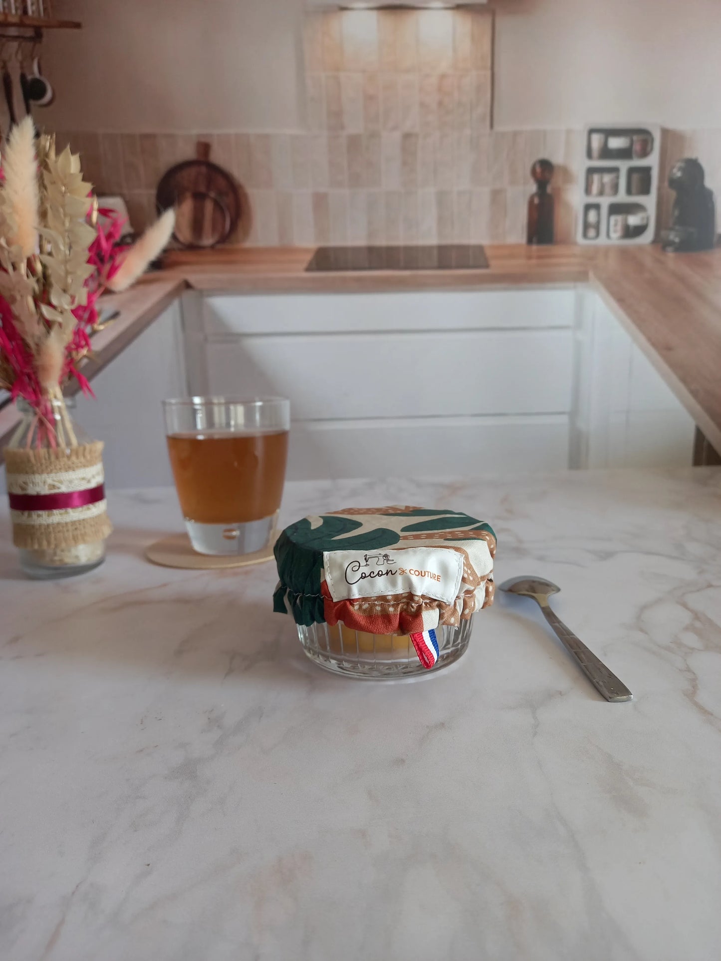 Charlotte alimentaire lavable à feuilles de palmier ajustée sur un petit ramequin en verre contenant une préparation jaune, avec l'étiquette Cocon Couture et un ruban tricolore visibles, sur un plan de travail en marbre près d'un verre de thé.