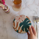 Couvre bol en tissu à feuilles de palmier vertes, terracotta et beige tenu d'une main au-dessus d'un plan de travail en marbre, près d'un verre de thé sur un dessous de verre et de couverts.