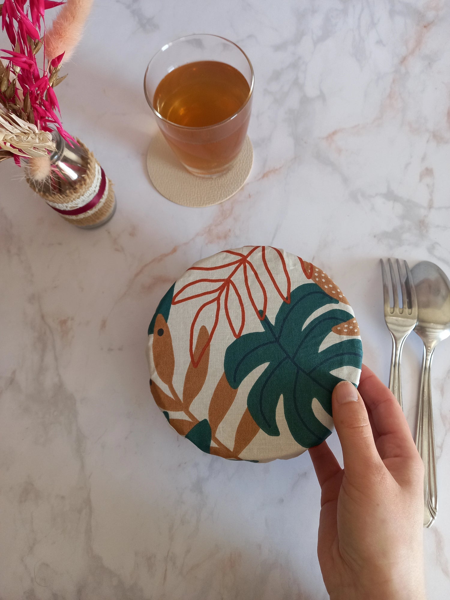 Couvre bol en tissu à feuilles de palmier vertes, terracotta et beige tenu d'une main au-dessus d'un plan de travail en marbre, près d'un verre de thé sur un dessous de verre et de couverts.
