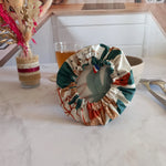 Couvre bol en tissu à feuilles de palmier retourné pour montrer son élastique froncé et son film alimentaire translucide intérieur, avec l'étiquette Cocon Couture et un ruban tricolore cousus au bord, posé près d'un bol beige.