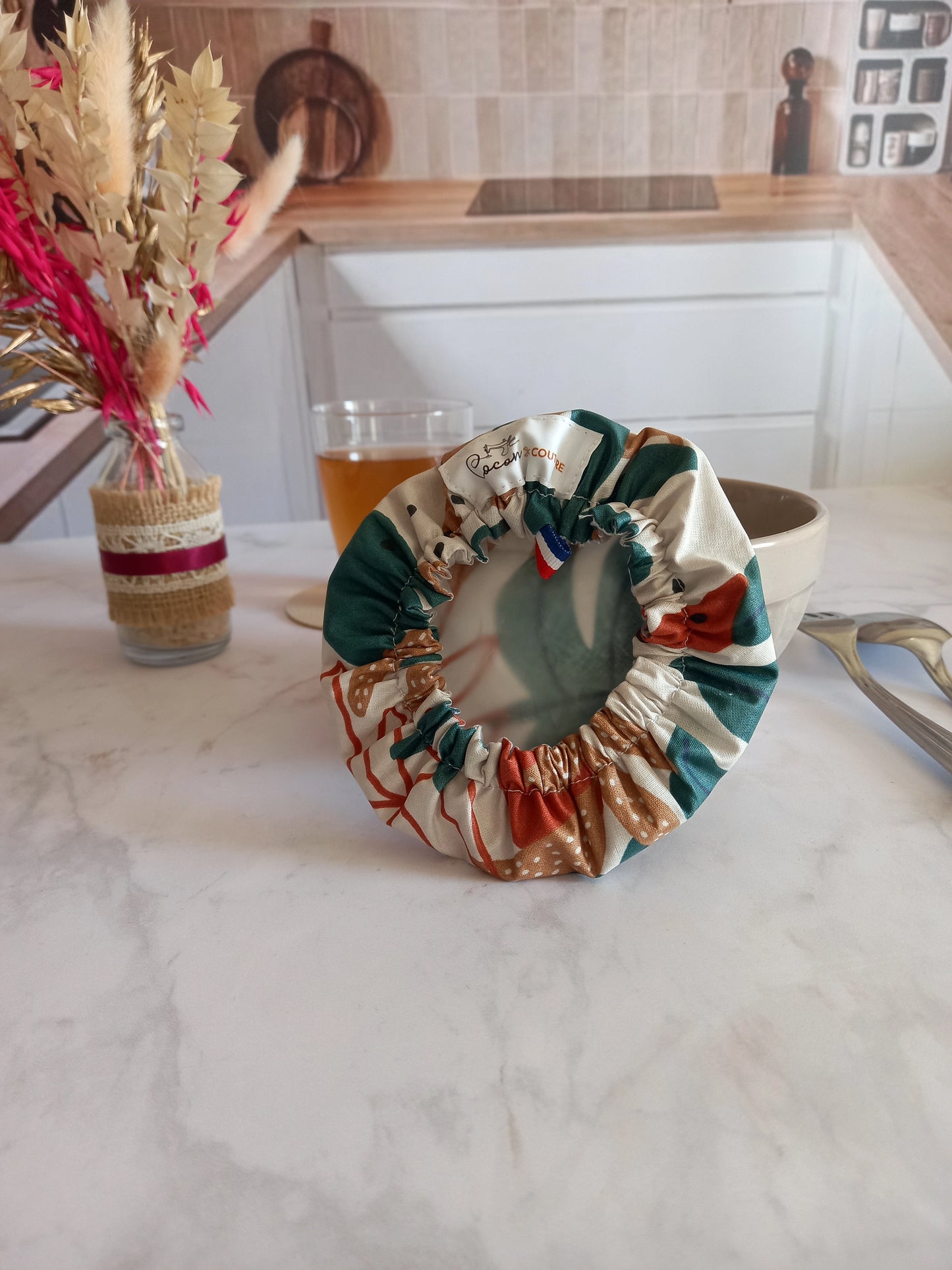 Couvre bol en tissu à feuilles de palmier retourné pour montrer son élastique froncé et son film alimentaire translucide intérieur, avec l'étiquette Cocon Couture et un ruban tricolore cousus au bord, posé près d'un bol beige.