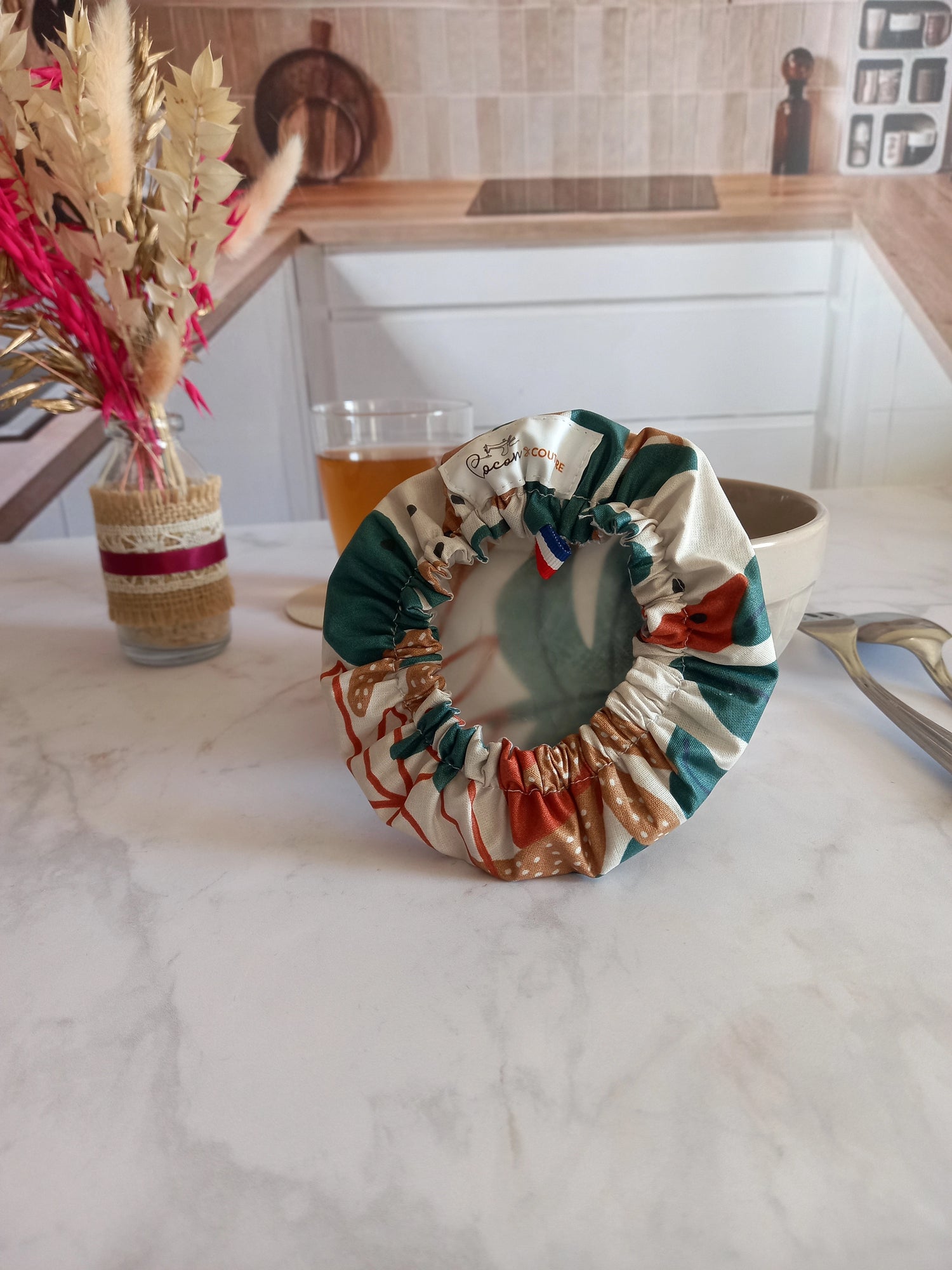 Couvre bol en tissu à feuilles de palmier retourné pour montrer son élastique froncé et son film alimentaire translucide intérieur, avec l'étiquette Cocon Couture et un ruban tricolore cousus au bord, posé près d'un bol beige.