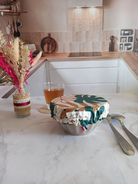 Couvre bol en tissu à feuilles de palmier ajusté sur un bol gris grâce à son élastique, avec l'étiquette Cocon Couture et un ruban tricolore visibles, sur un plan de travail en marbre près d'un verre de thé.
