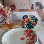 Assiette à fleurs rouges posée au premier plan dans une cuisine, derrière laquelle un couvre saladier en tissu à feuillage tropical recouvre un saladier, près d'un verre de thé et d'un bouquet de fleurs séchées.