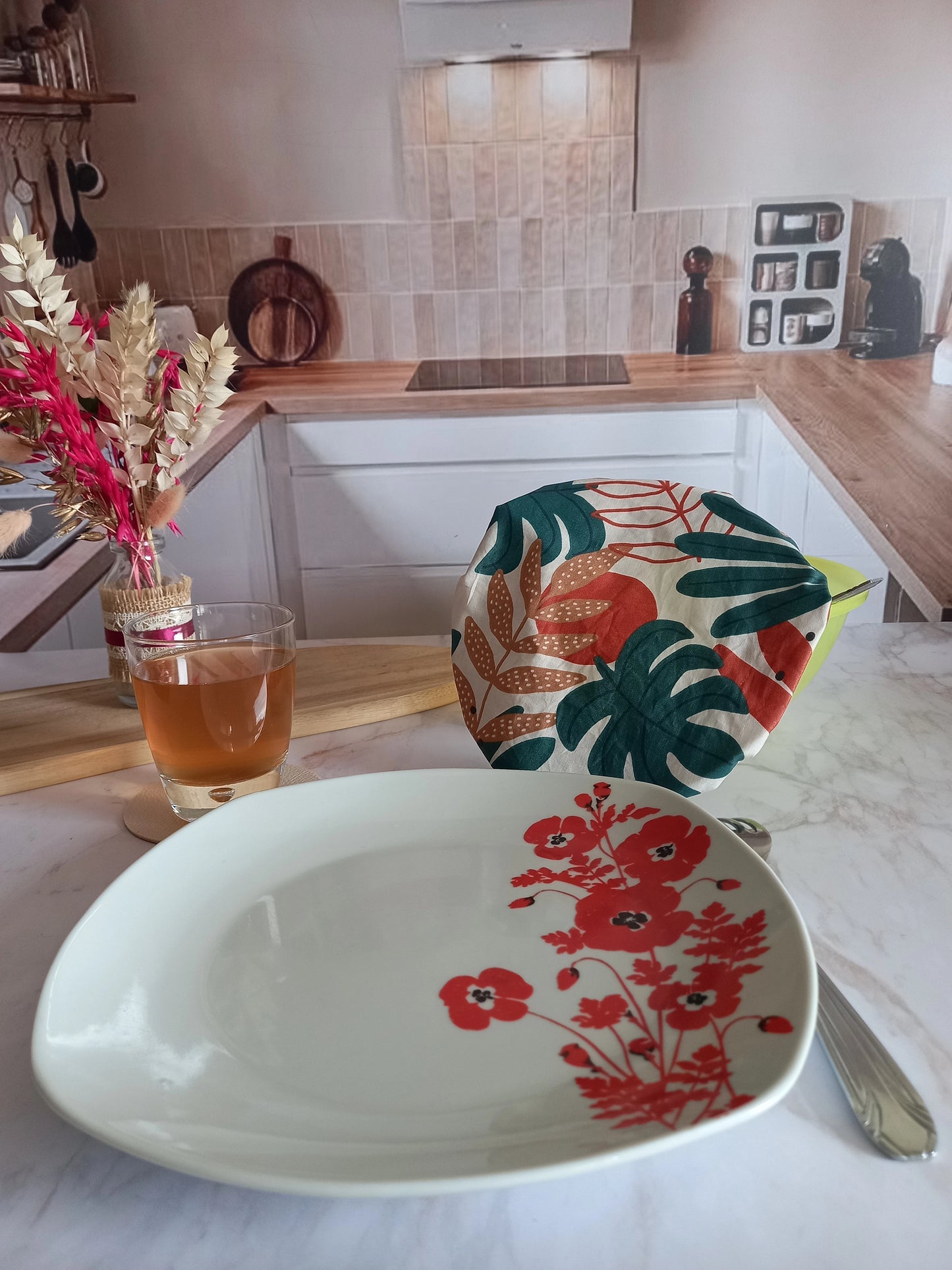 Assiette à fleurs rouges posée au premier plan dans une cuisine, derrière laquelle un couvre saladier en tissu à feuillage tropical recouvre un saladier, près d'un verre de thé et d'un bouquet de fleurs séchées.