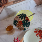 Couvre saladier en tissu à feuillage tropical vert, terracotta et beige posé sur un saladier jaune avec une cuillère, sur un plan de travail en marbre où se trouvent un verre de thé et une assiette à fleurs rouges.