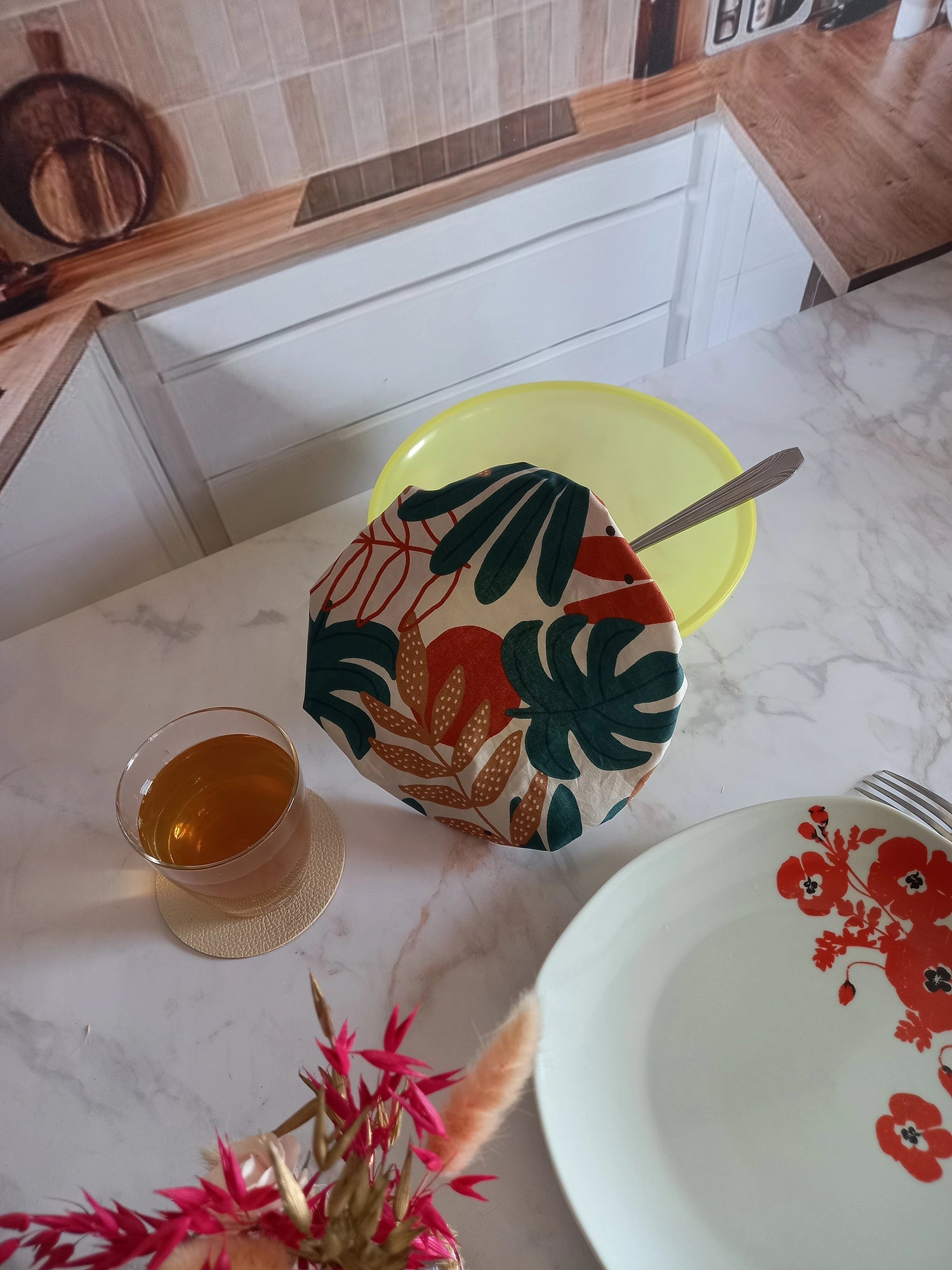 Couvre saladier en tissu à feuillage tropical vert, terracotta et beige posé sur un saladier jaune avec une cuillère, sur un plan de travail en marbre où se trouvent un verre de thé et une assiette à fleurs rouges.