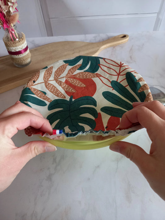 Deux mains ajustent l'élastique d'un couvre saladier en tissu à feuillage tropical sur le bord d'un saladier vert, le ruban tricolore bleu blanc rouge cousu au bord restant visible.