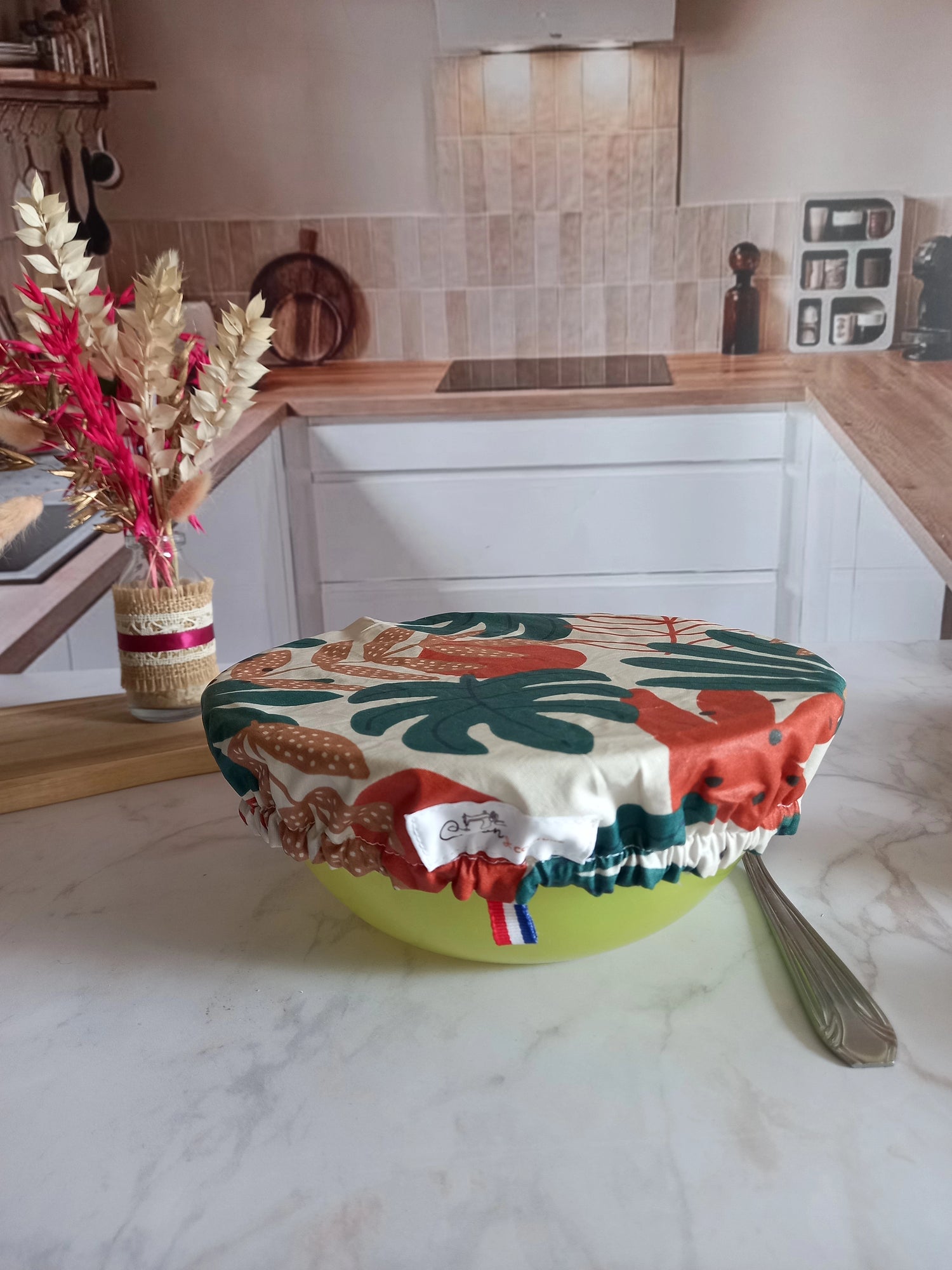 Couvre saladier en tissu à feuillage tropical enfilé sur un saladier vert anis grâce à son élastique froncé, avec une étiquette de marque cousue et un ruban tricolore sur le pourtour, sur un plan de travail en marbre.