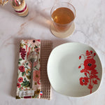 La feuille essuie-tout lavable au motif floral rose et bordeaux est pliée sur un plan de travail en marbre, une cuillère posée dessus. À côté se trouvent une assiette à fleurs rouges et un verre de thé.