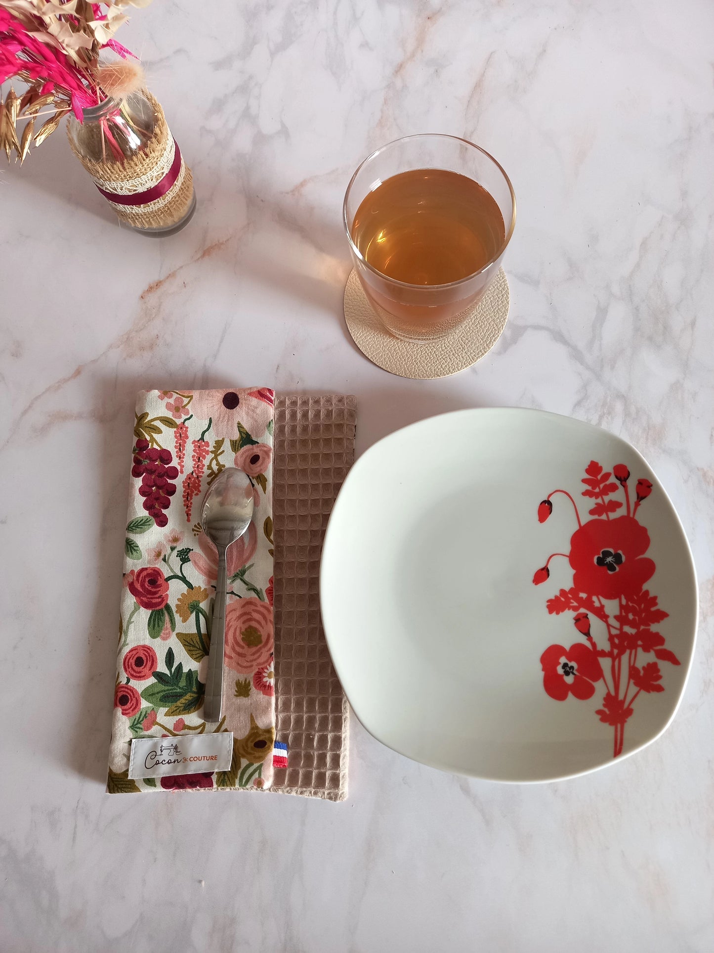 La feuille essuie-tout lavable au motif floral rose et bordeaux est pliée sur un plan de travail en marbre, une cuillère posée dessus. À côté se trouvent une assiette à fleurs rouges et un verre de thé.