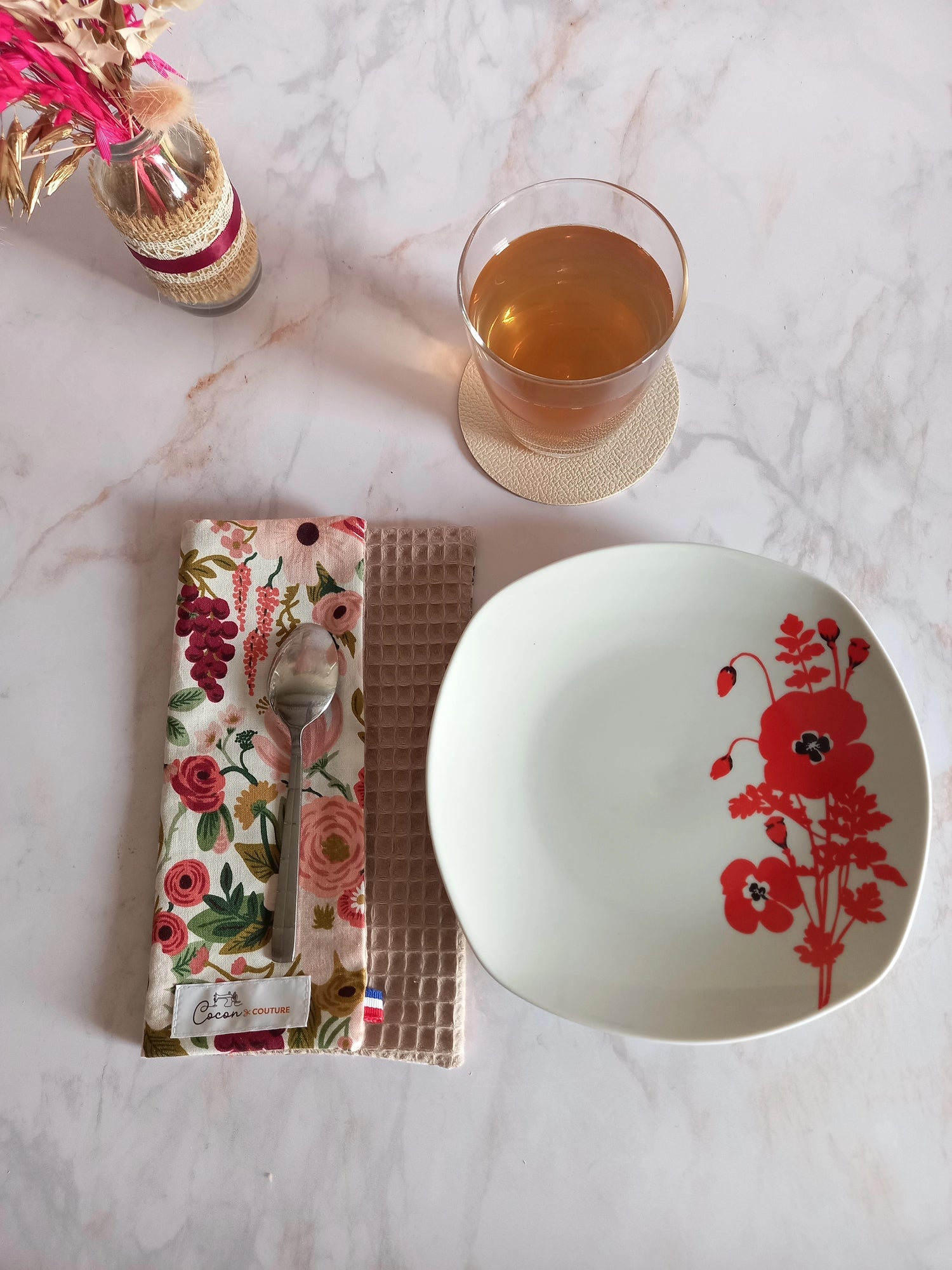 La feuille essuie-tout lavable au motif floral rose et bordeaux est pliée sur un plan de travail en marbre, une cuillère posée dessus. À côté se trouvent une assiette à fleurs rouges et un verre de thé.