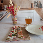 La feuille essuie-tout lavable au motif floral rose et bordeaux est pliée sur un plan de travail en marbre, dans une cuisine. Elle est posée près d'une assiette et d'un verre de thé.