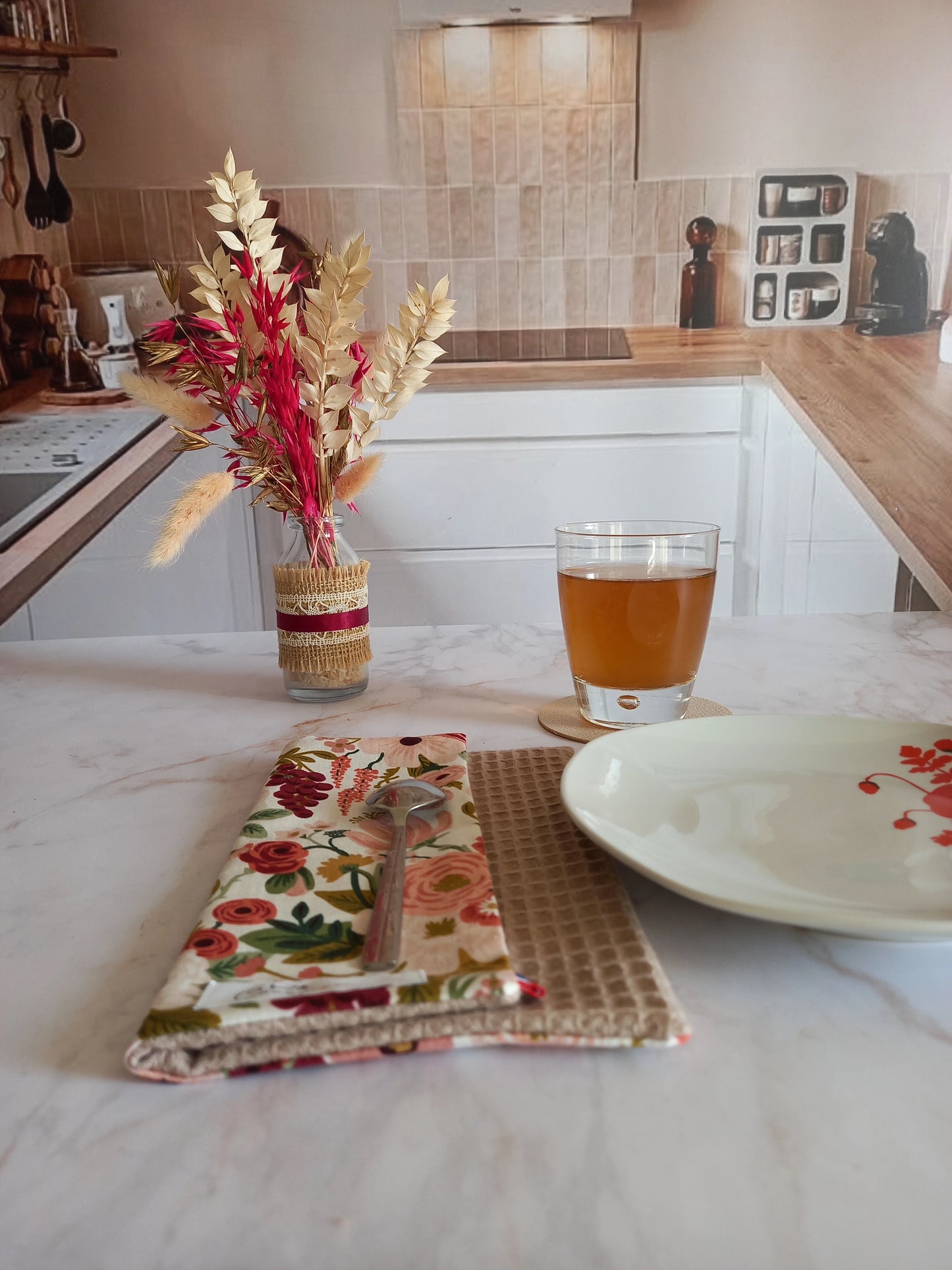 La feuille essuie-tout lavable au motif floral rose et bordeaux est pliée sur un plan de travail en marbre, dans une cuisine. Elle est posée près d'une assiette et d'un verre de thé.