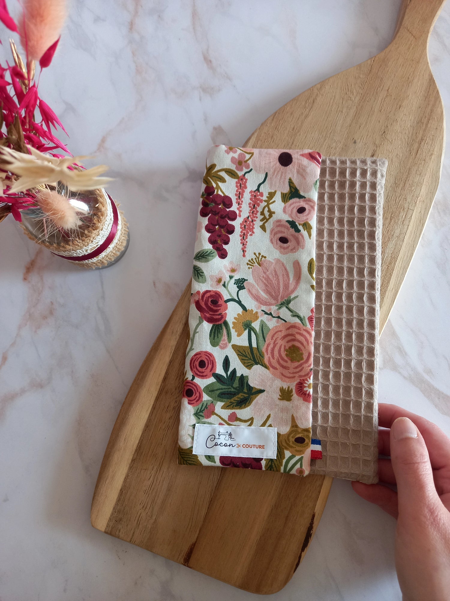 La feuille essuie-tout lavable est pliée sur une planche en bois. On aperçoit son recto au motif floral rose et bordeaux, son envers en nid d'abeille beige et l'étiquette Cocon Couture.