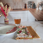 La feuille essuie-tout lavable au motif floral rose et doré est pliée sur un plan de travail en marbre, dans une cuisine. Une cuillère est posée dessus, près d'une assiette à fleurs rouges et d'un verre de thé.