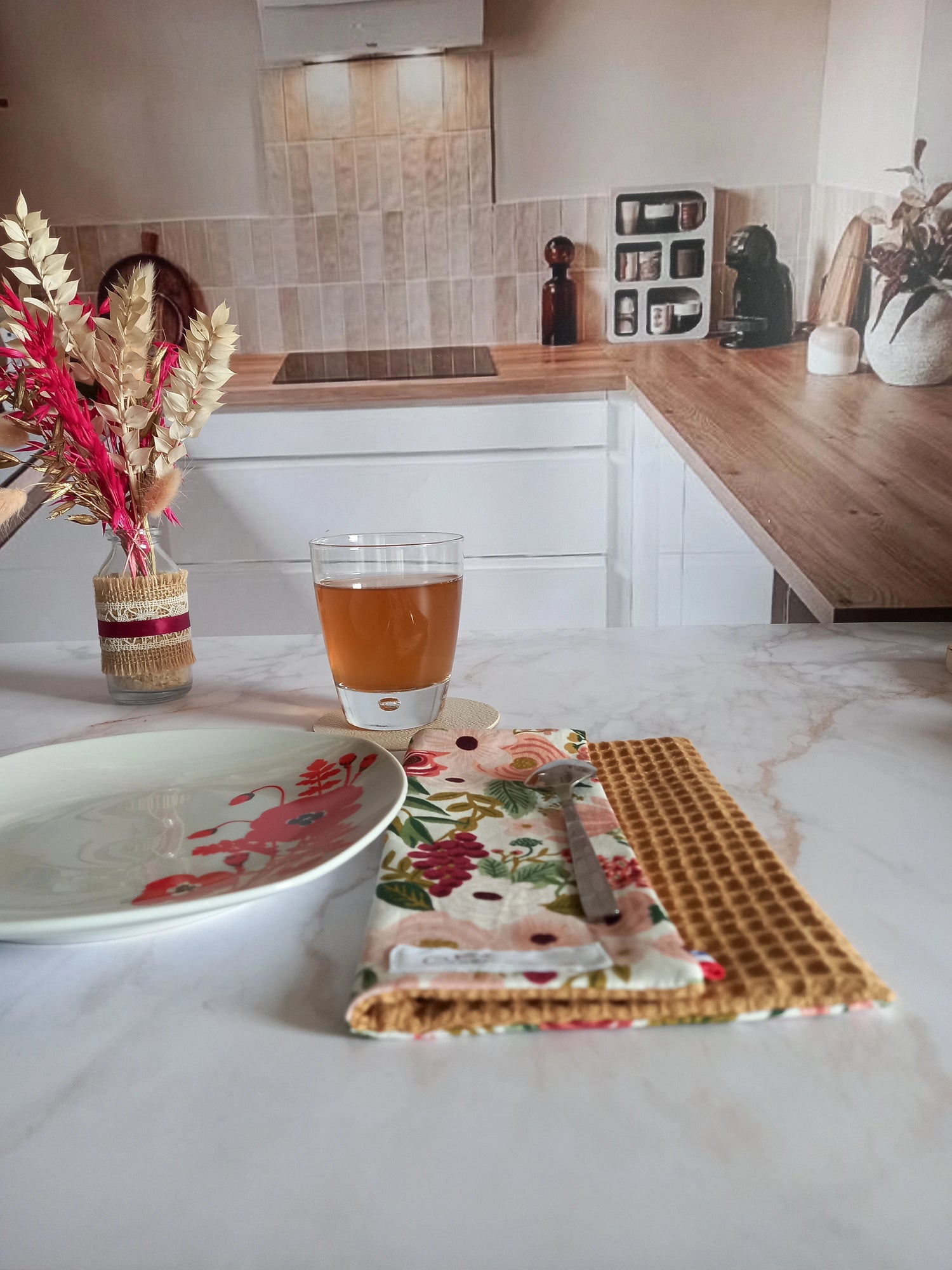 La feuille essuie-tout lavable au motif floral rose et doré est pliée sur un plan de travail en marbre, dans une cuisine. Une cuillère est posée dessus, près d'une assiette à fleurs rouges et d'un verre de thé.
