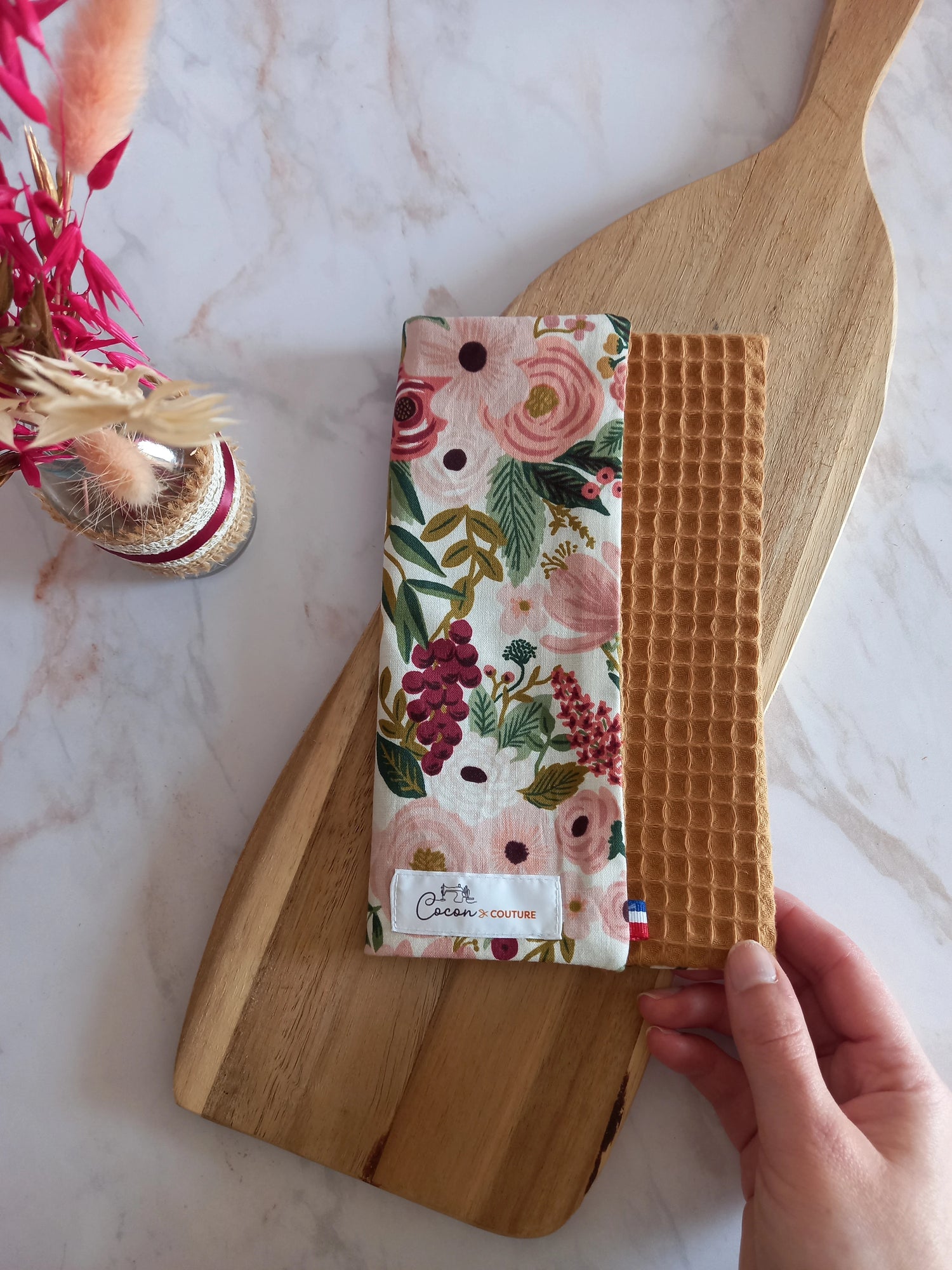 La feuille essuie-tout lavable est pliée sur une planche en bois. On aperçoit son recto au motif floral rose et doré, son envers en nid d'abeille caramel et l'étiquette Cocon Couture.