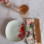 La feuille essuie-tout lavable au motif floral rose et doré est pliée sur un plan de travail en marbre, vue de dessus. Elle côtoie une assiette à fleurs rouges, un verre de thé et l'étiquette Cocon Couture.