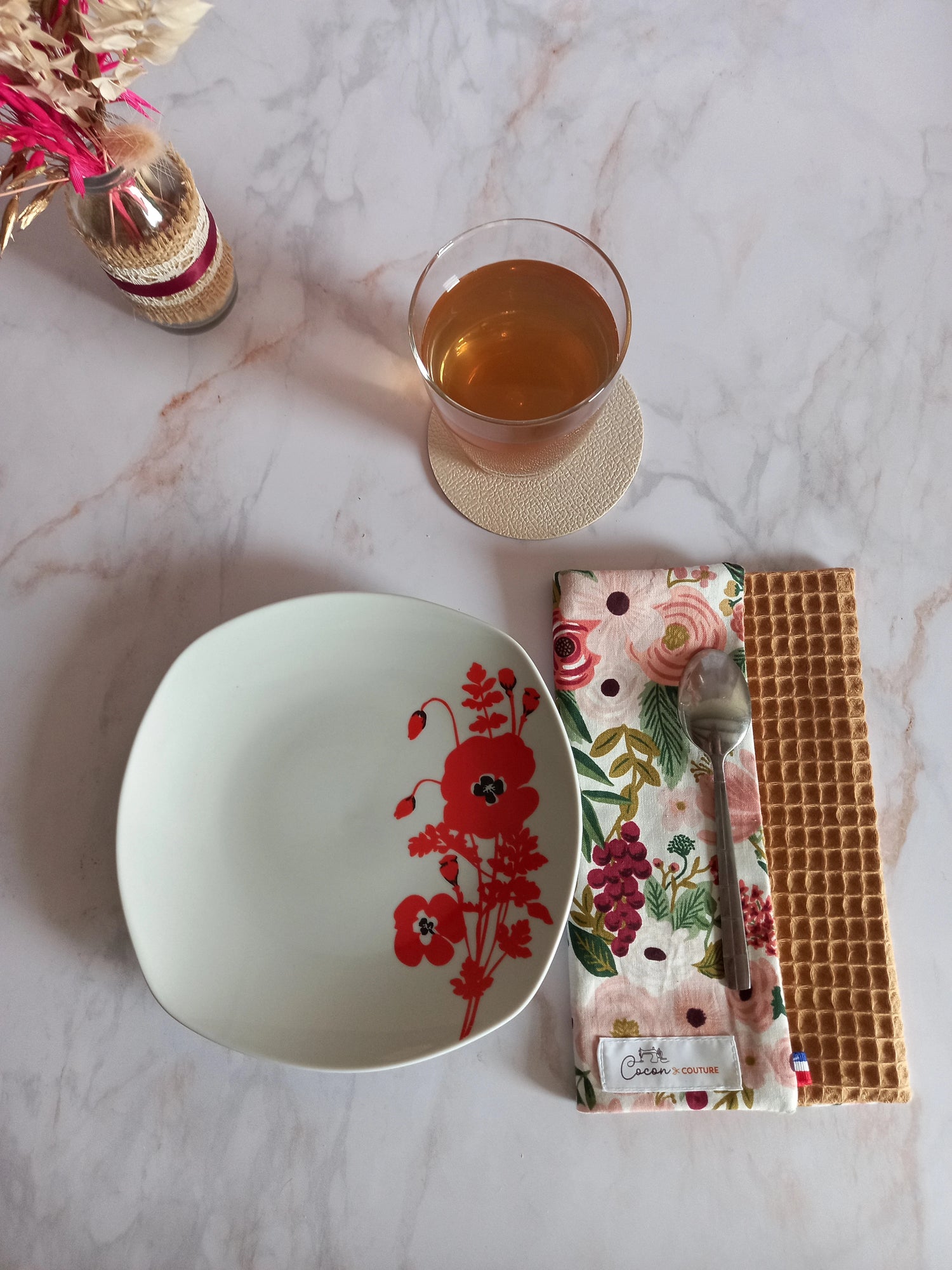 La feuille essuie-tout lavable au motif floral rose et doré est pliée sur un plan de travail en marbre, vue de dessus. Elle côtoie une assiette à fleurs rouges, un verre de thé et l'étiquette Cocon Couture.