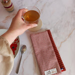 La feuille essuie-tout lavable à carreaux vichy terracotta est pliée sur un plan de travail en marbre, vue de dessus. Elle est posée près d'une assiette à fleurs rouges et d'une cuillère, une main tenant un verre de thé.