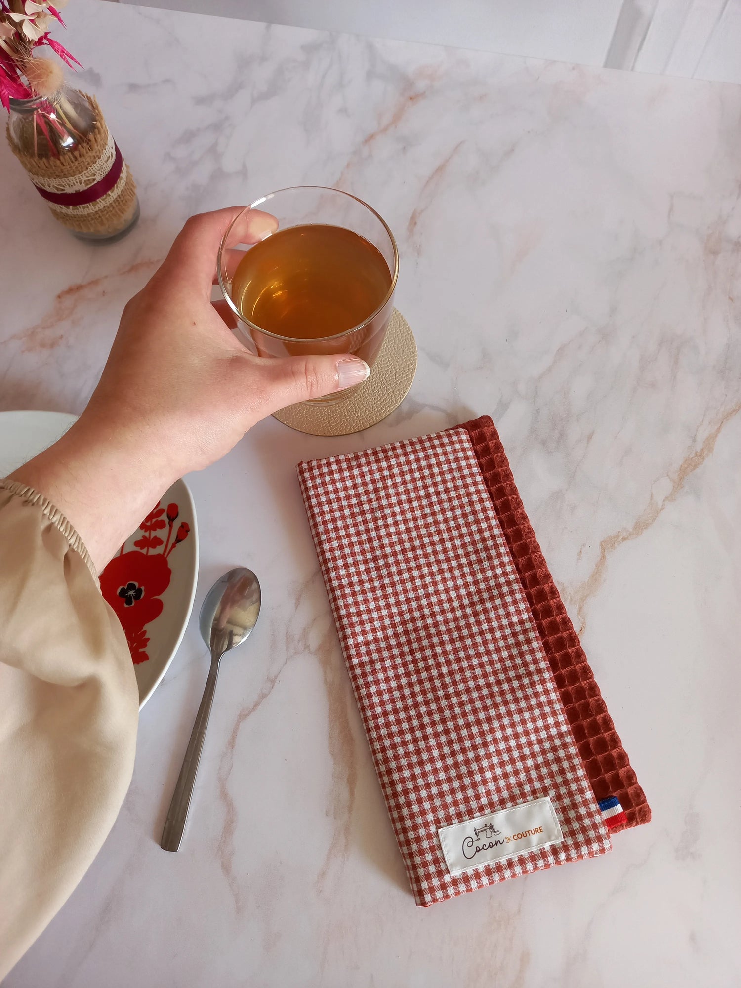 La feuille essuie-tout lavable à carreaux vichy terracotta est pliée sur un plan de travail en marbre, vue de dessus. Elle est posée près d'une assiette à fleurs rouges et d'une cuillère, une main tenant un verre de thé.