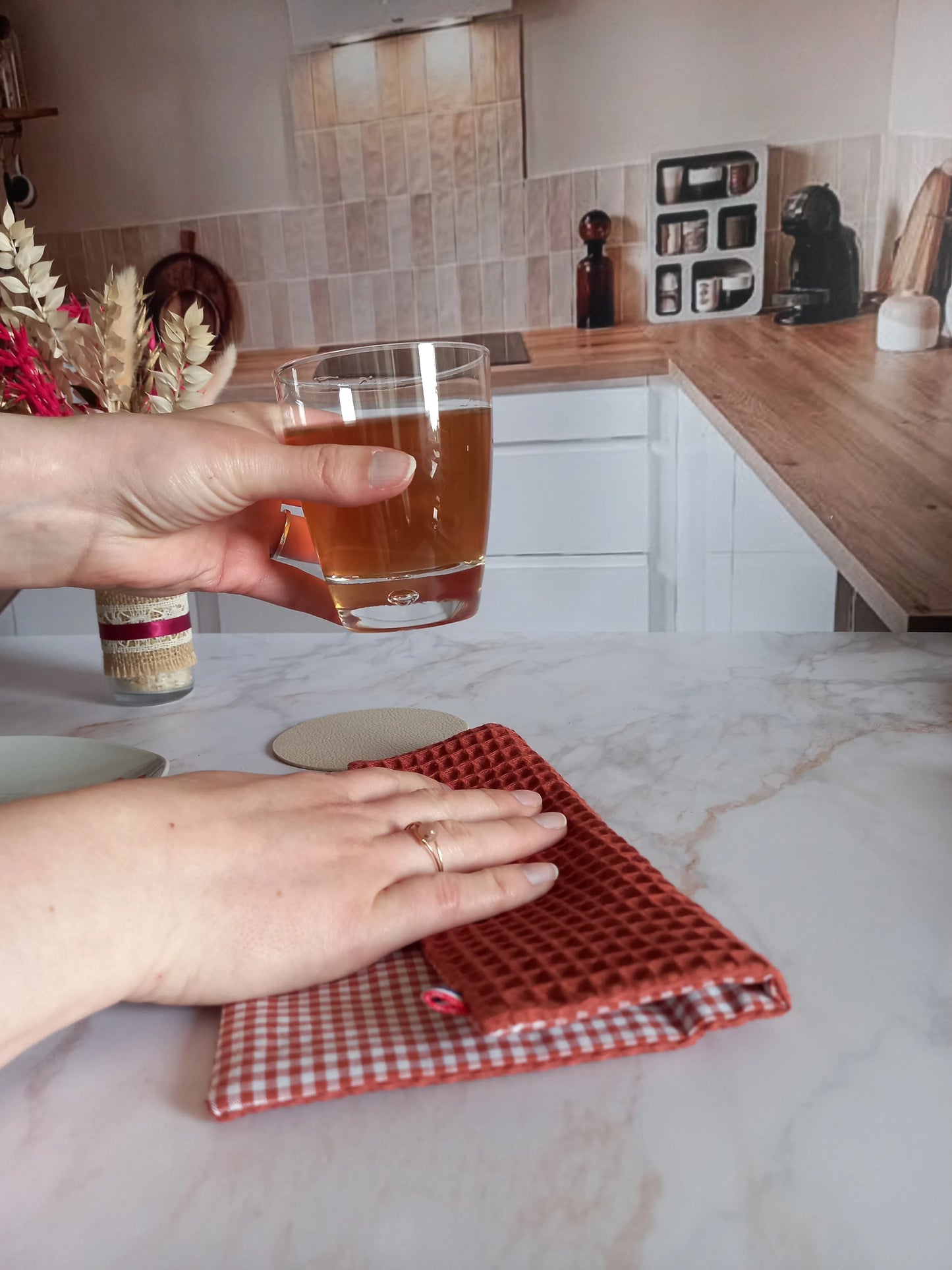 La feuille essuie-tout lavable, côté nid d'abeille terracotta, est pliée sur un plan de travail en marbre. Une main est posée dessus tandis que l'autre tient un verre de thé, dans une cuisine.