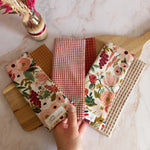 Trois feuilles essuie-tout lavables des collections dune fleurie, jardin doré et nos souvenirs sont disposées en éventail sur une planche en bois et tenues par une main. Chacune associe un côté motif ou carreaux à un envers en nid d'abeille, et les étiquettes Cocon Couture sont visibles.