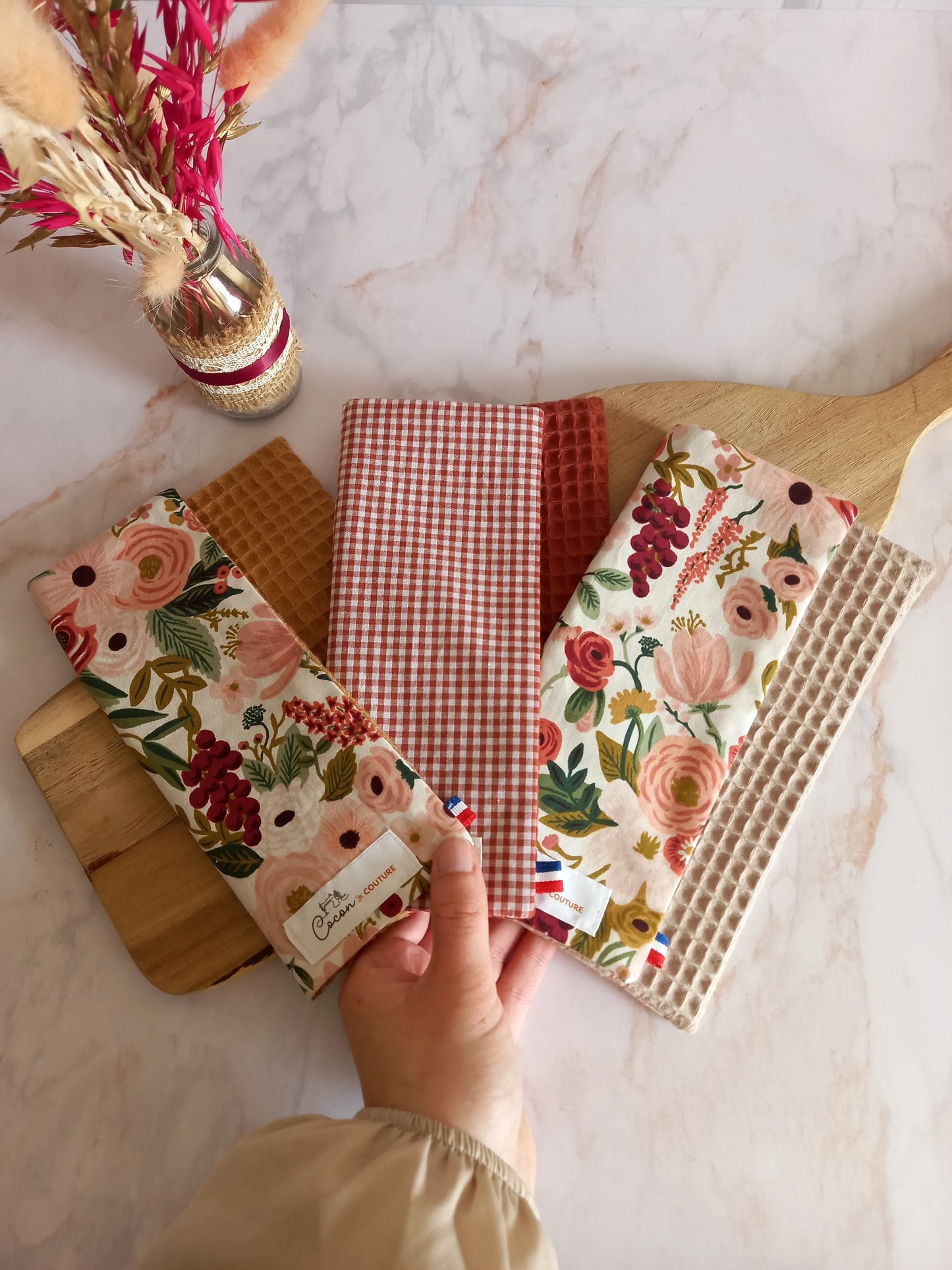 Trois feuilles essuie-tout lavables des collections dune fleurie, jardin doré et nos souvenirs sont disposées en éventail sur une planche en bois et tenues par une main. Chacune associe un côté motif ou carreaux à un envers en nid d'abeille, et les étiquettes Cocon Couture sont visibles.