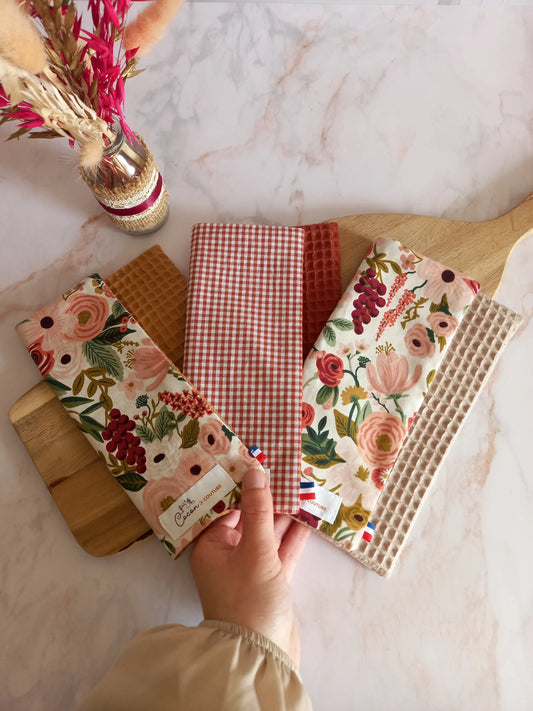 Trois feuilles essuie-tout lavables des collections dune fleurie, jardin doré et nos souvenirs sont disposées en éventail sur une planche en bois et tenues par une main. Chacune associe un côté motif ou carreaux à un envers en nid d'abeille, et les étiquettes Cocon Couture sont visibles.
