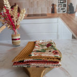 Trois feuilles essuie-tout lavables des collections dune fleurie, jardin doré et nos souvenirs sont empilées et pliées sur une planche en bois, dans une cuisine. On distingue leurs faces en nid d'abeille moutarde, terracotta et beige, et un côté à motif floral rose.