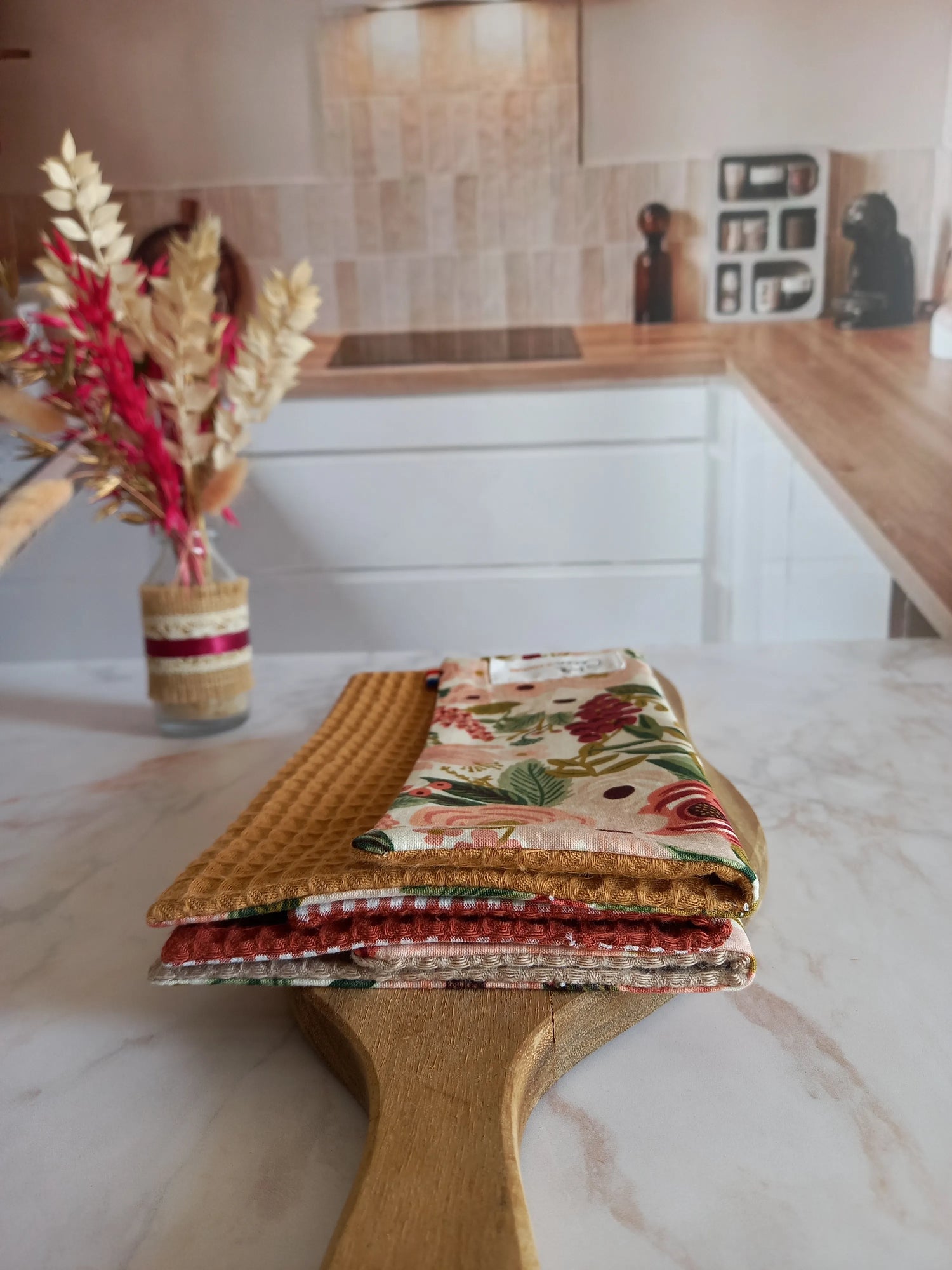 Trois feuilles essuie-tout lavables des collections dune fleurie, jardin doré et nos souvenirs sont empilées et pliées sur une planche en bois, dans une cuisine. On distingue leurs faces en nid d'abeille moutarde, terracotta et beige, et un côté à motif floral rose.