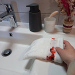 Gant démaquillant lavable en tissu lisse blanc et bande à coquelicots rouges, enfilé sur une main inclinée vers la vasque, dans une salle de bain.