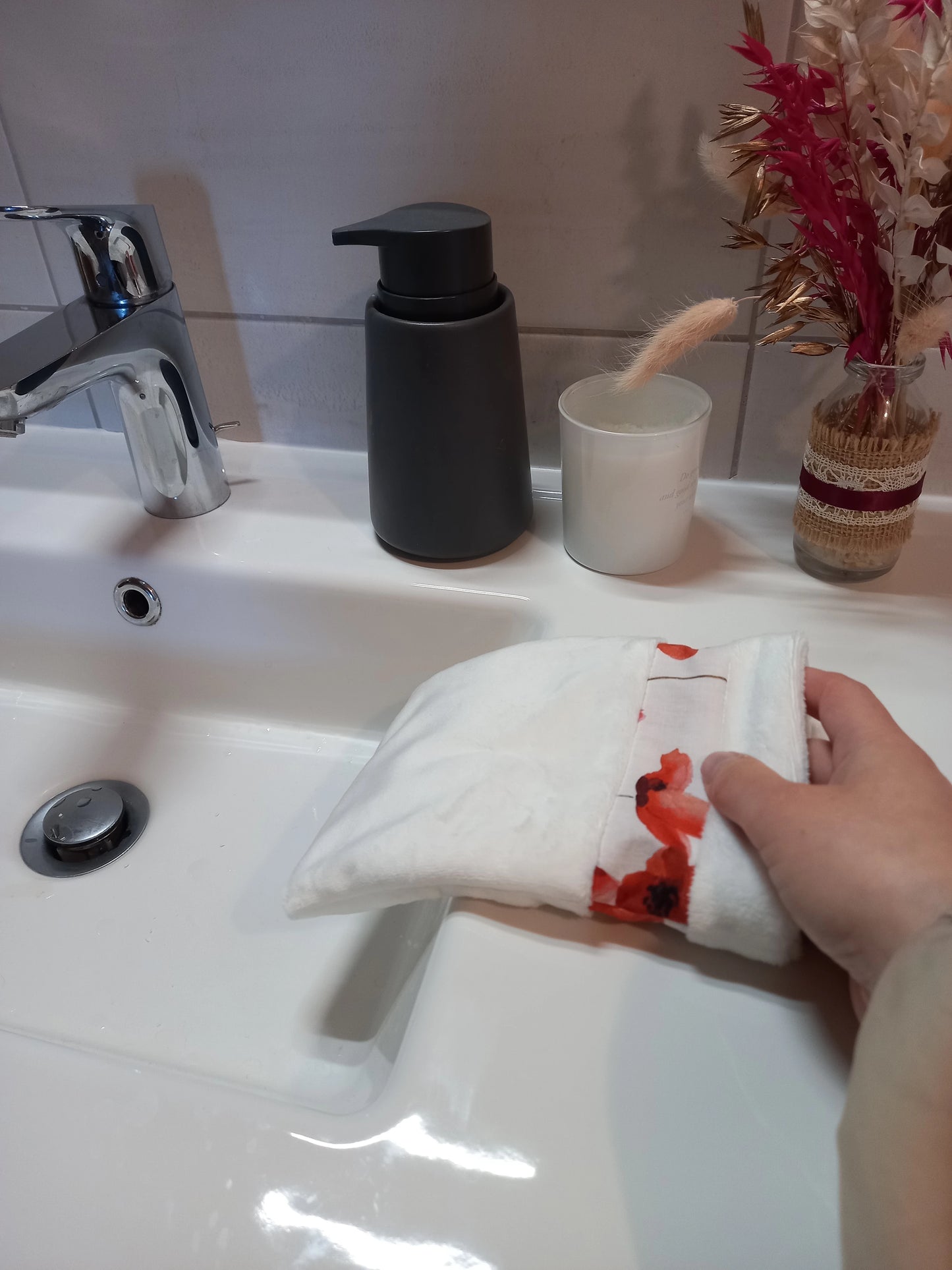 Gant démaquillant lavable en tissu lisse blanc et bande à coquelicots rouges, enfilé sur une main inclinée vers la vasque, dans une salle de bain.