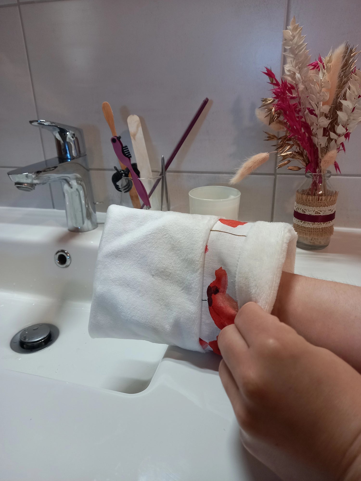Main portant un gant démaquillant lavable en tissu lisse blanc avec sa bande à coquelicots rouges, au-dessus du lavabo.