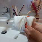 Gant démaquillant lavable en tissu lisse blanc, orné d'une bande à coquelicots rouges, enfilé sur une main au-dessus d'un lavabo.