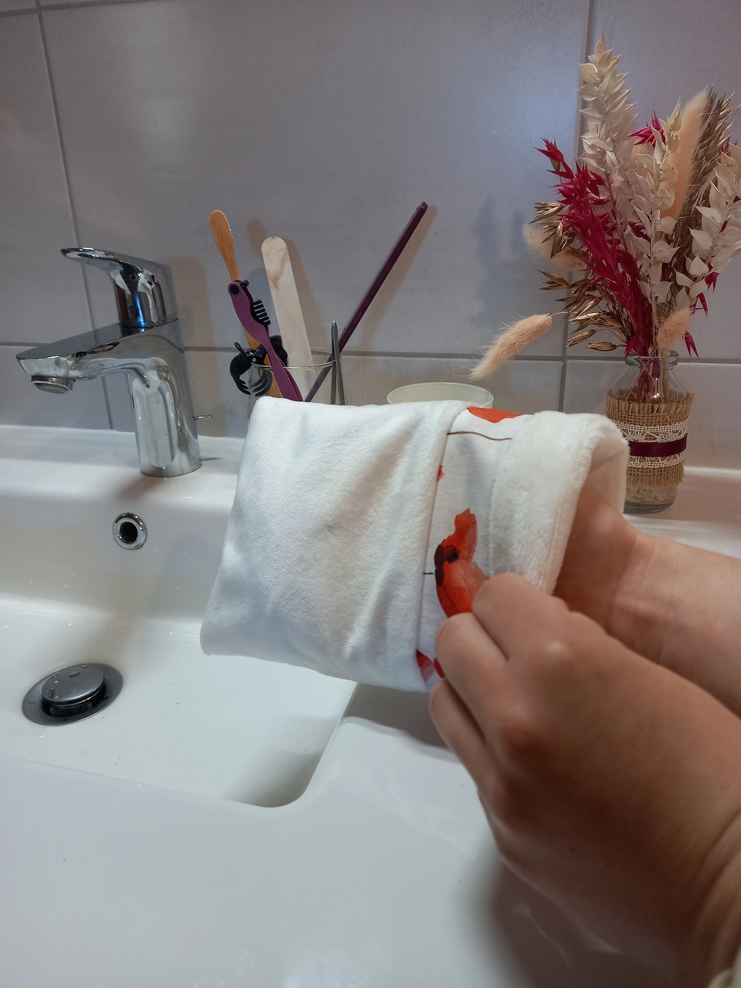 Gant démaquillant lavable en tissu lisse blanc, orné d'une bande à coquelicots rouges, enfilé sur une main au-dessus d'un lavabo.