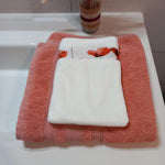 Gant démaquillant lavable en tissu lisse blanc, orné d'une bande à coquelicots rouges, posé à plat sur une serviette éponge corail.