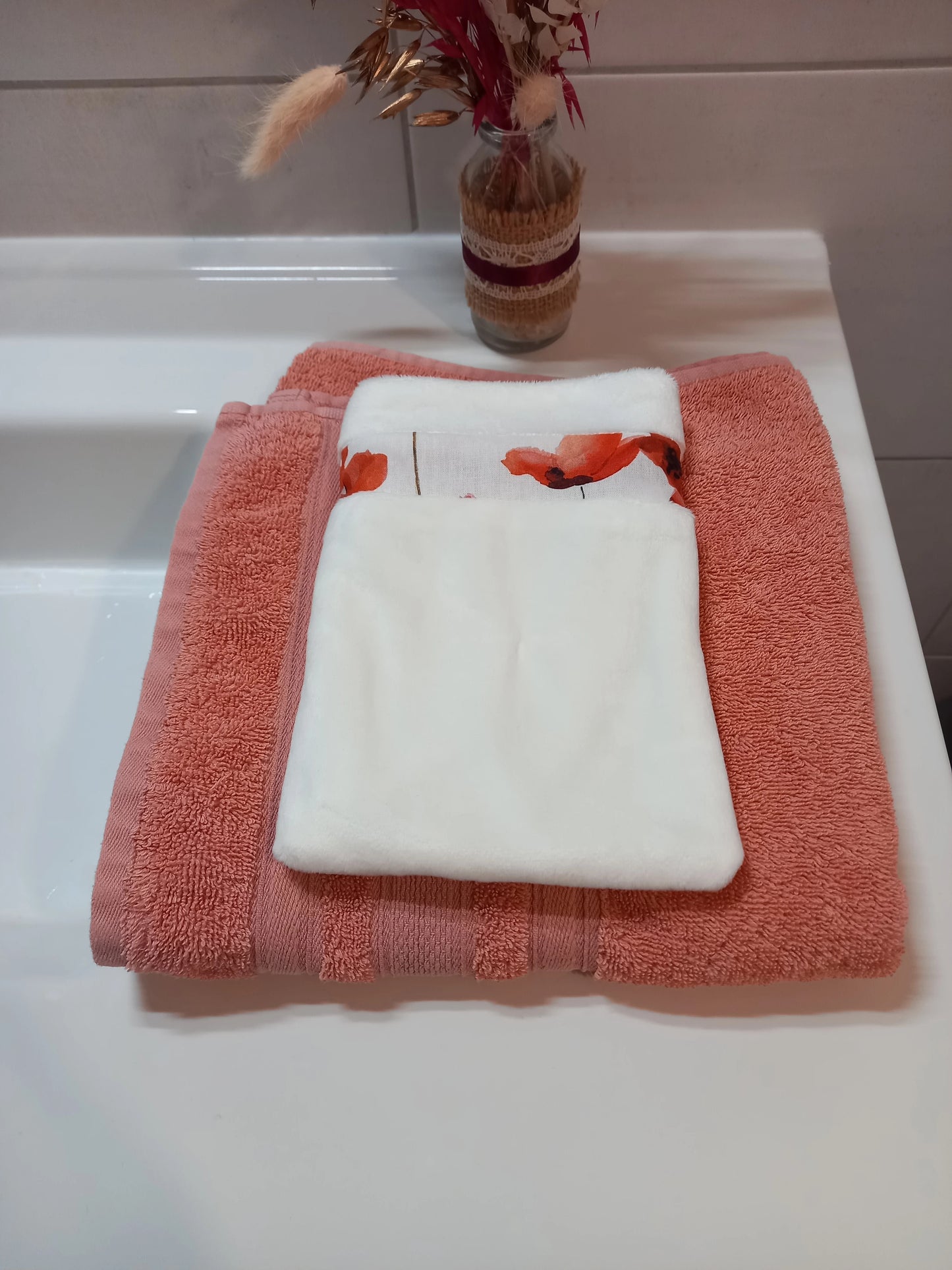 Gant démaquillant lavable en tissu lisse blanc, orné d'une bande à coquelicots rouges, posé à plat sur une serviette éponge corail.