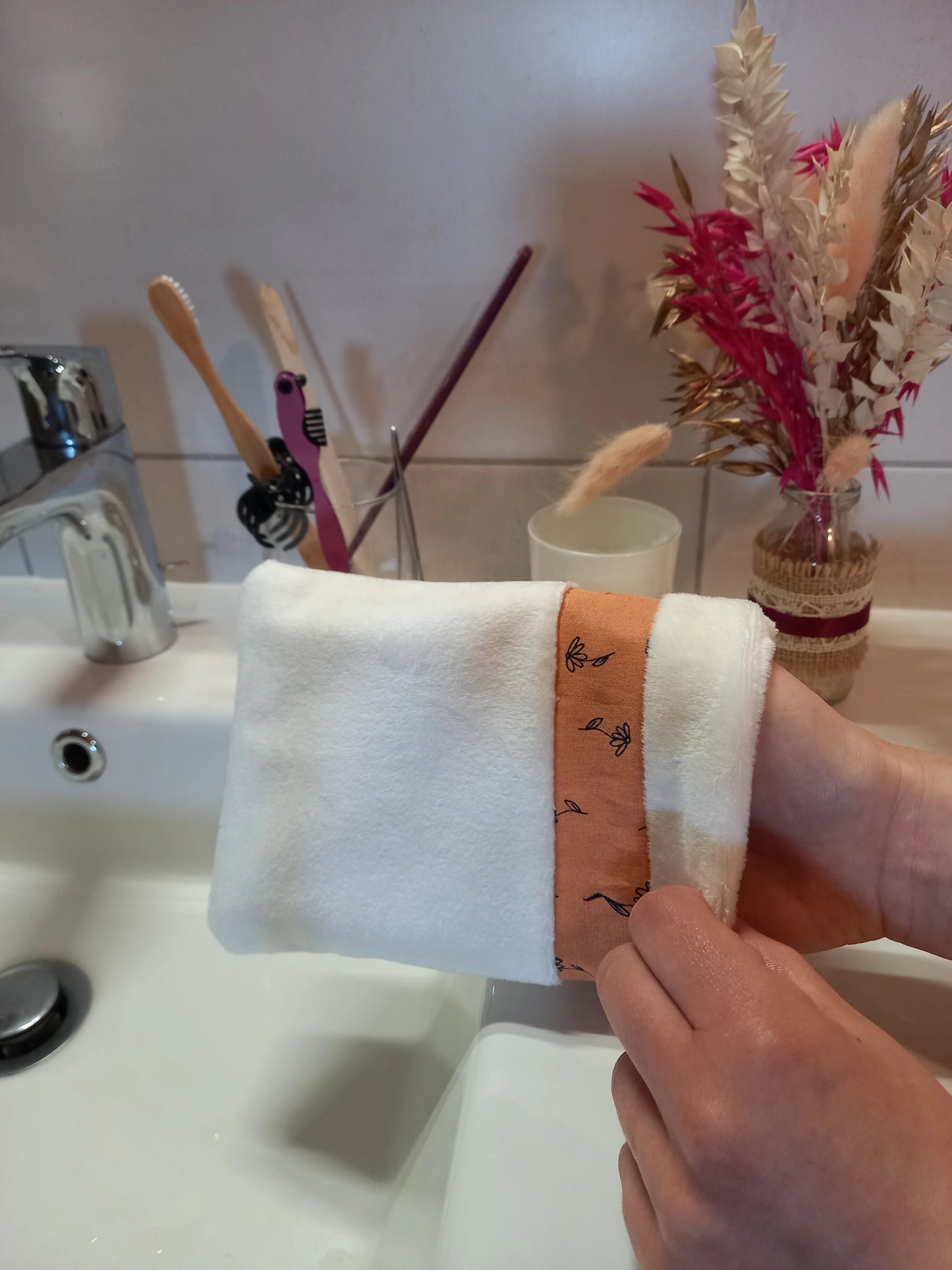 Gant démaquillant lavable en velours lisse blanc à bande fleurs printanières orange, porté sur la main avec le poignet visible, devant un lavabo de salle de bain et un bouquet de fleurs séchées.