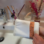 Gant démaquillant lavable en velours lisse blanc, orné d'une bande à fleurs printanières orange, enfilé sur une main au-dessus d'un lavabo de salle de bain, près d'un verre à brosses à dents et d'un bouquet de fleurs séchées.