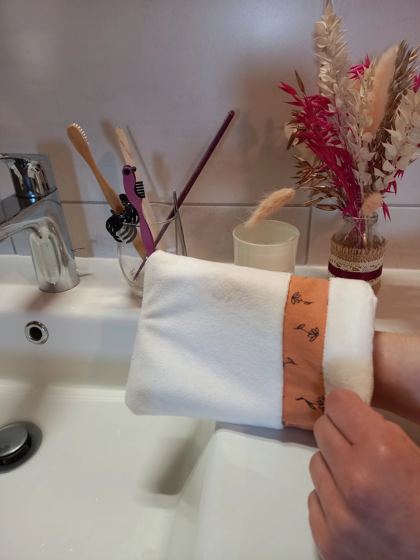 Gant démaquillant lavable en velours lisse blanc, orné d'une bande à fleurs printanières orange, enfilé sur une main au-dessus d'un lavabo de salle de bain, près d'un verre à brosses à dents et d'un bouquet de fleurs séchées.