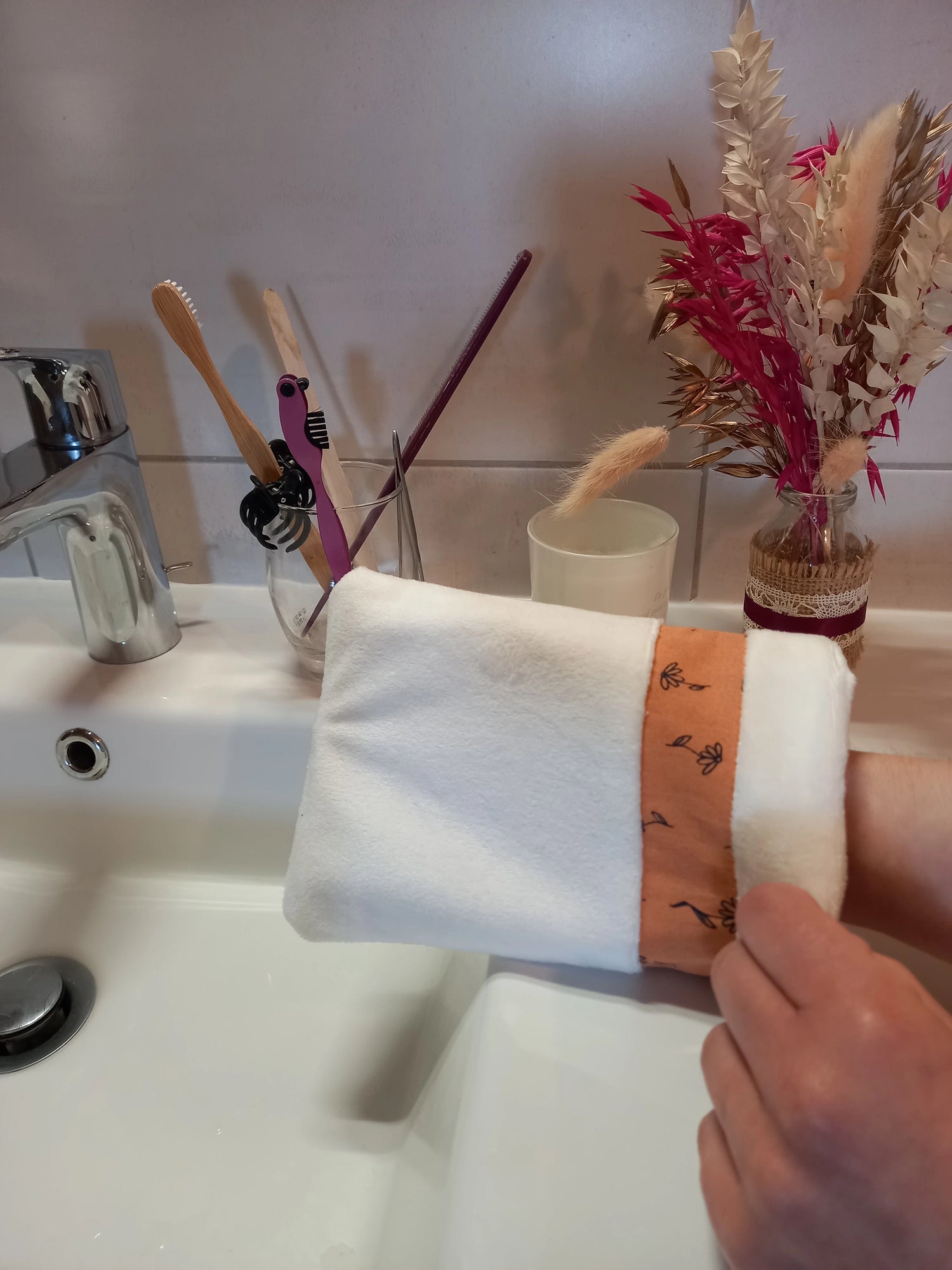 Gant démaquillant lavable en velours lisse blanc, orné d'une bande à fleurs printanières orange, enfilé sur une main au-dessus d'un lavabo de salle de bain, près d'un verre à brosses à dents et d'un bouquet de fleurs séchées.