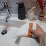 Gant démaquillant lavable en tissu lisse blanc, orné d'une bande à fleurs printanières orange, enfilé sur une main au-dessus d'un lavabo.