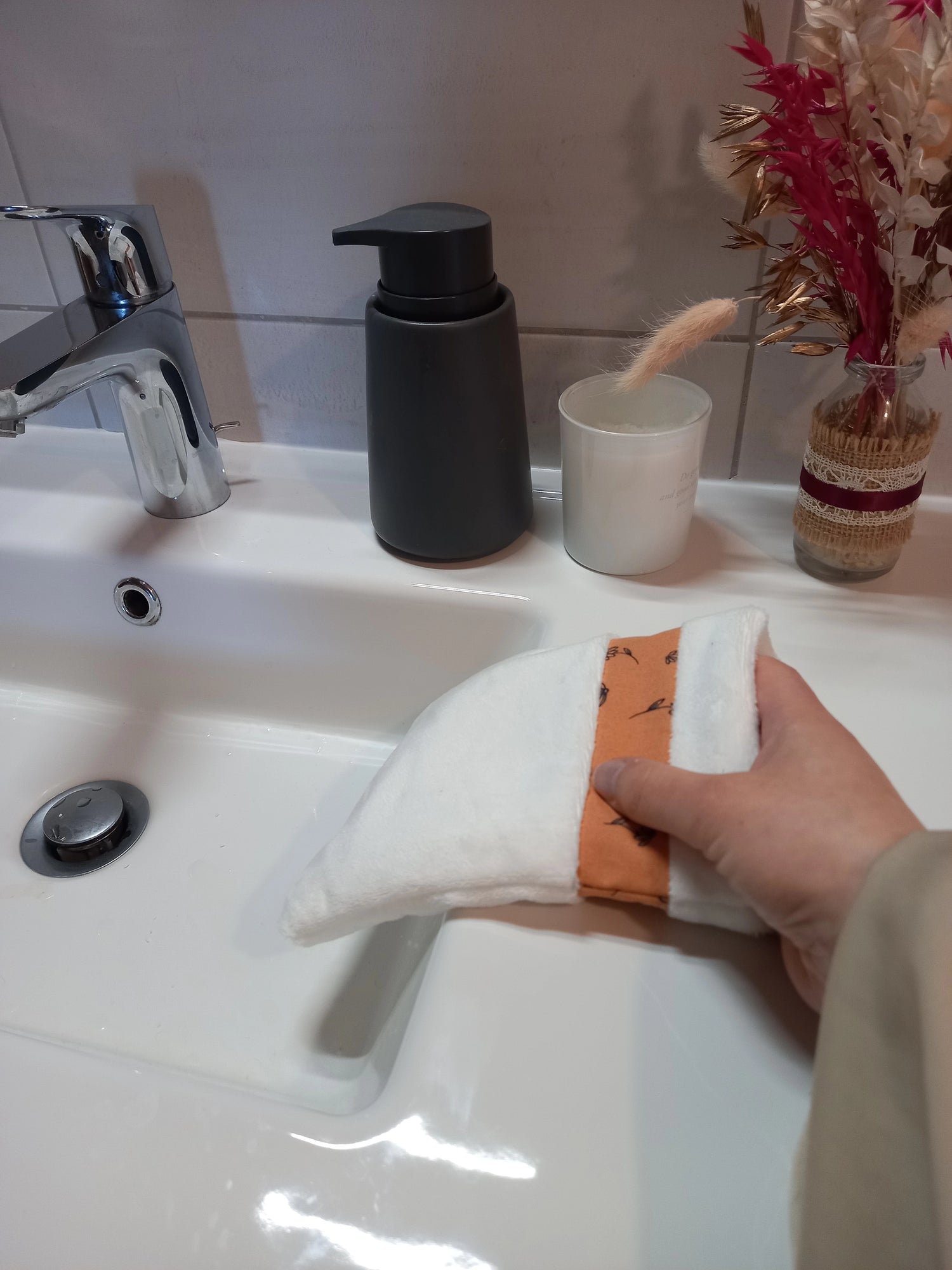 Gant démaquillant lavable en tissu lisse blanc, orné d'une bande à fleurs printanières orange, enfilé sur une main au-dessus d'un lavabo.