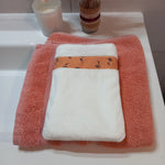 Gant démaquillant lavable en tissu lisse blanc, orné d'une bande à fleurs printanières orange, posé à plat sur une serviette éponge corail.