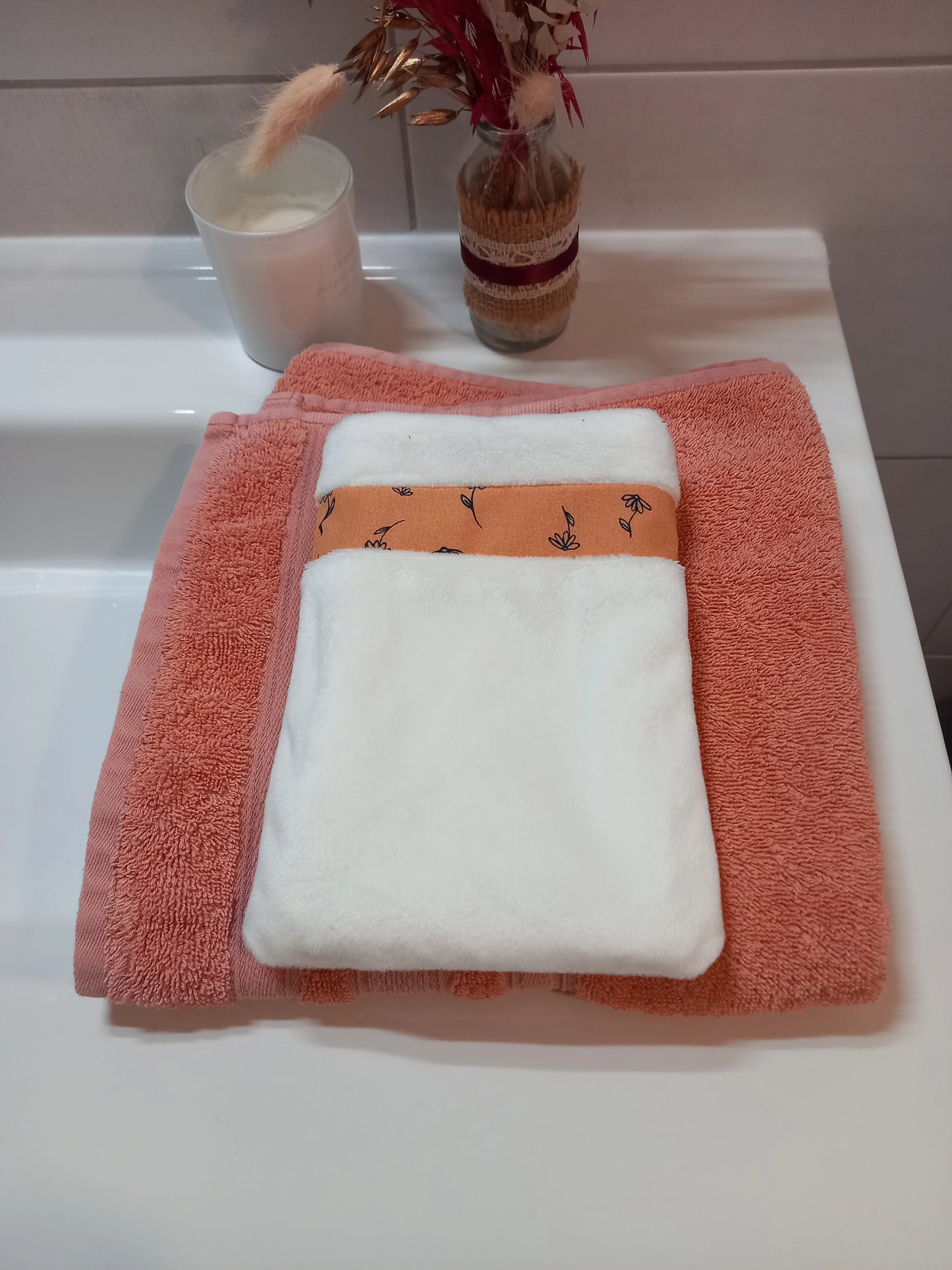 Gant démaquillant lavable en tissu lisse blanc, orné d'une bande à fleurs printanières orange, posé à plat sur une serviette éponge corail.