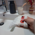 Gant démaquillant lavable en minky blanc à pois en relief et bande à coquelicots rouges, enfilé sur une main inclinée vers la vasque, dans une salle de bain.